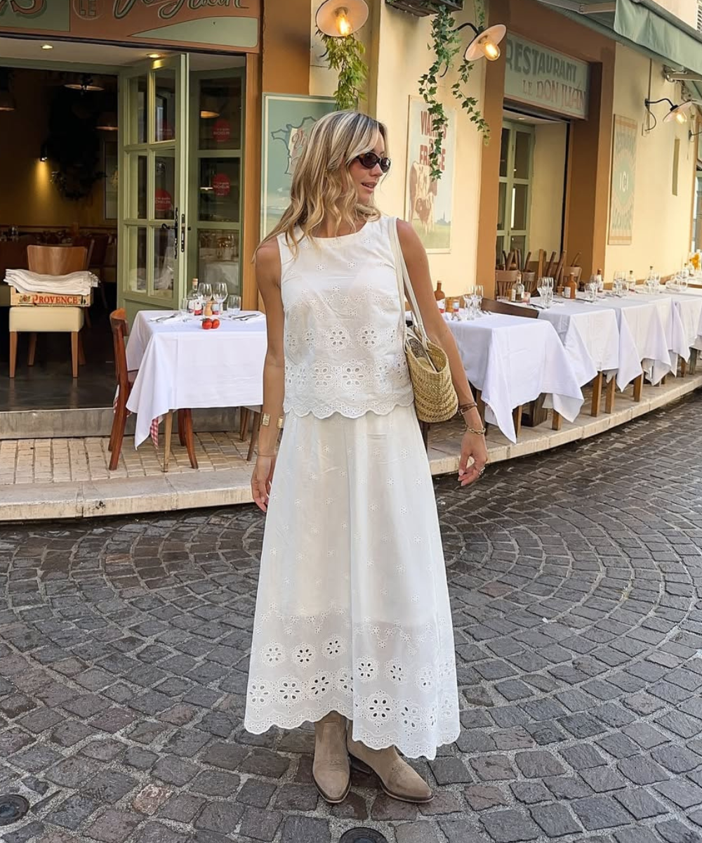 Modelo caminha na rua com saia rendada branca midi de tecido leve, combinada com blusa sem mangas e botas bege, transmitindo