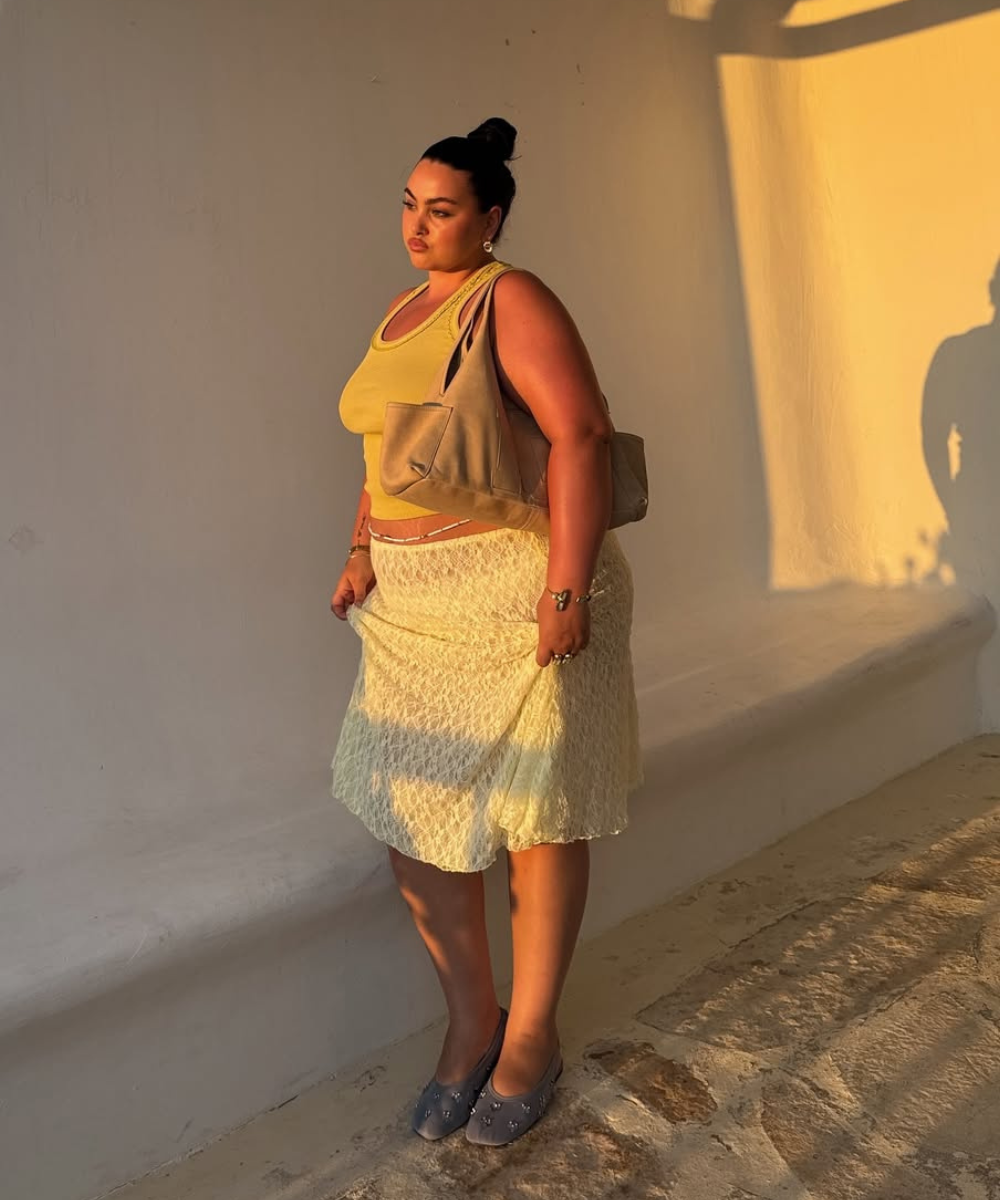 Modelo posa em luz dourada usando saia rendada midi off-white, regata amarela e bolsa bege, transmitindo elegância casual.
