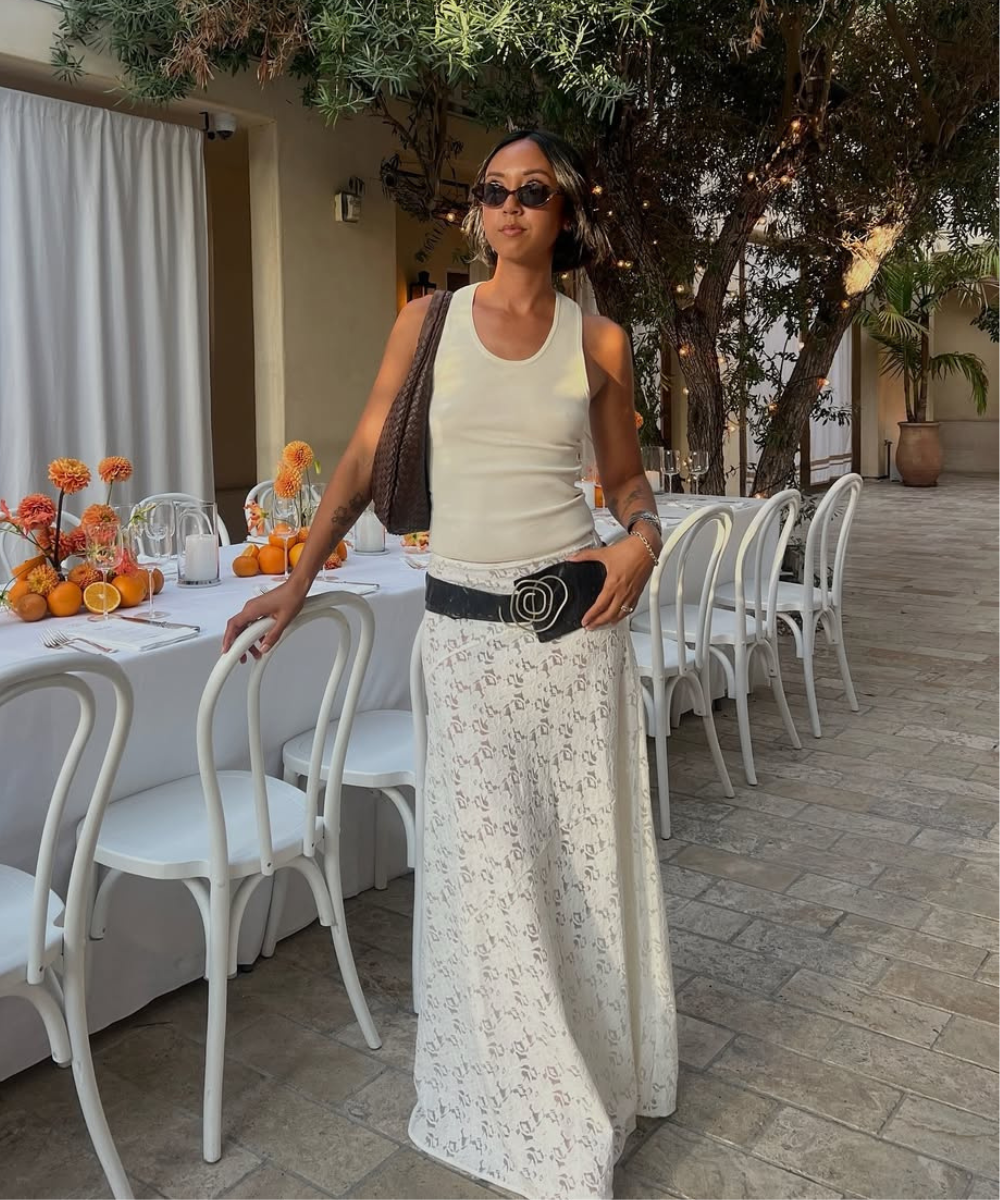 Modelo posa com saia rendada branca longa, regata off-white e acessórios em preto, transmitindo elegância casual em ambiente