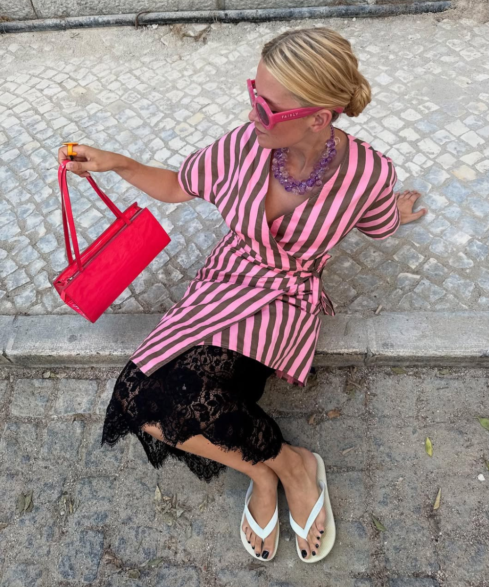 Mulher sentada com saia rendada preta midi, blusa rosa com listras marrons, bolsa vermelha e chinelos brancos, estilo casual
