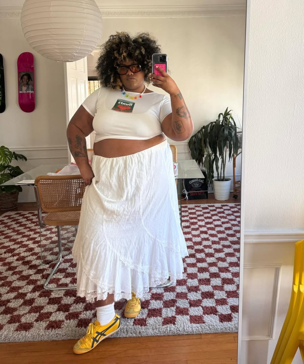 Modelo posa em ambiente interno com saia rendada branca fluida, cropped branco, tênis amarelo e acessórios coloridos,