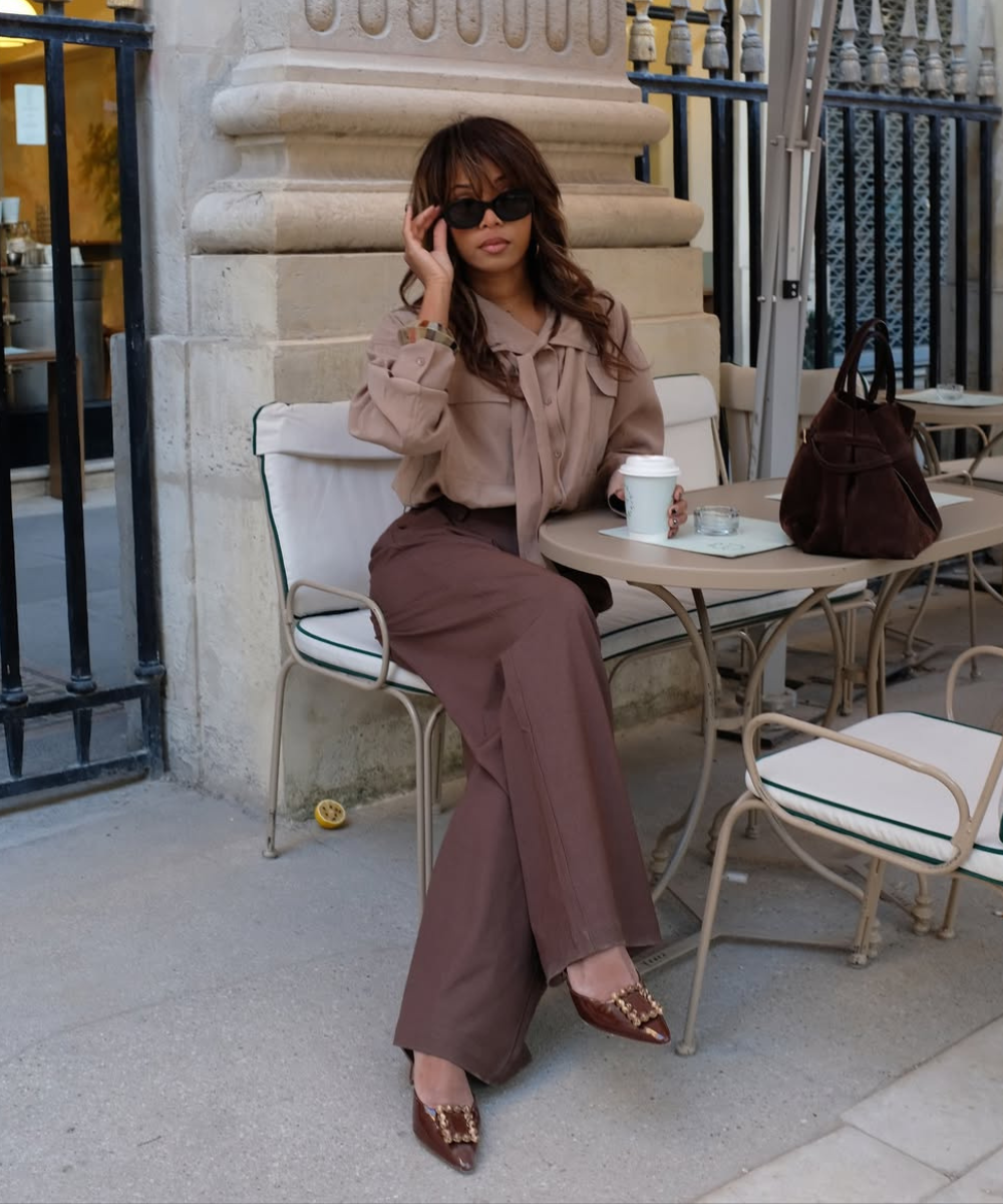 Look esporte fino com blusa nude de laço e calça marrom. Acessórios incluem óculos escuros e bolsa marrom. Estilo elegante.