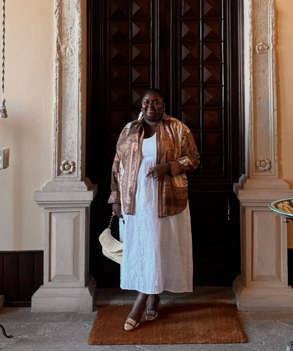 Modelo posa confiante com vestido branco de linho fluido e camisa estampada em tons terrosos, criando look esporte fino