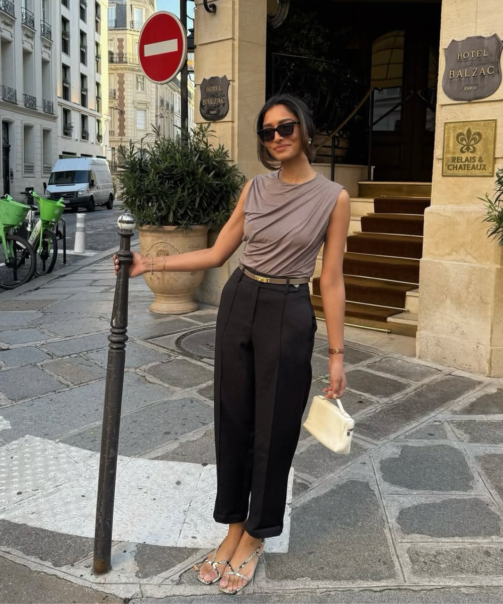 Look esporte fino com blusa drapeada em tom neutro e calça preta de corte reto. Acessórios como bolsa branca e sandálias com detalhes em strass complementam o visual elegante. A pose confiante realça a sofisticação, ideal para eventos sociais.