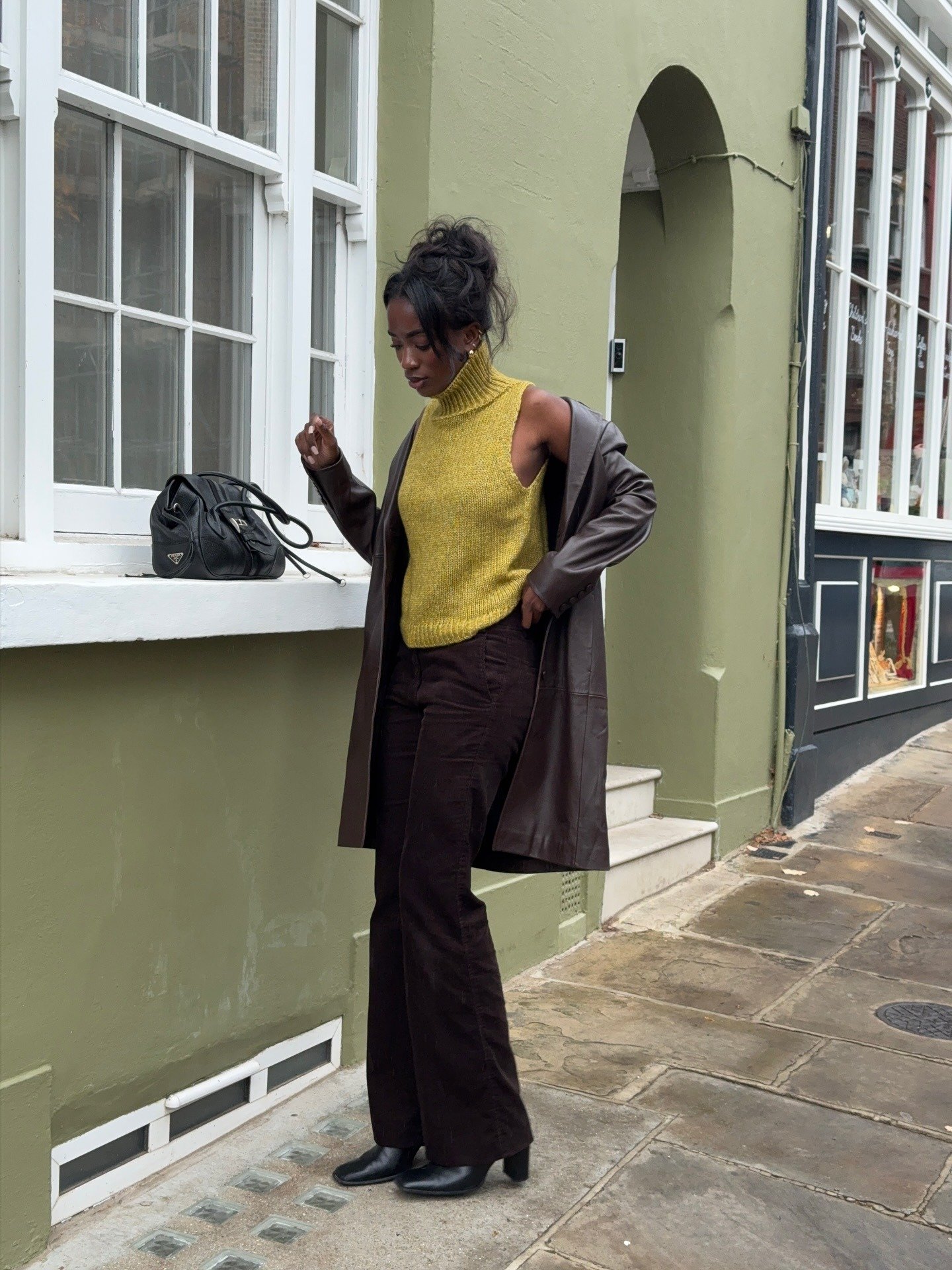 Modelo posa com look natal verde composto por suéter de tricô sem mangas, gola alta, calça flare marrom e sobretudo de