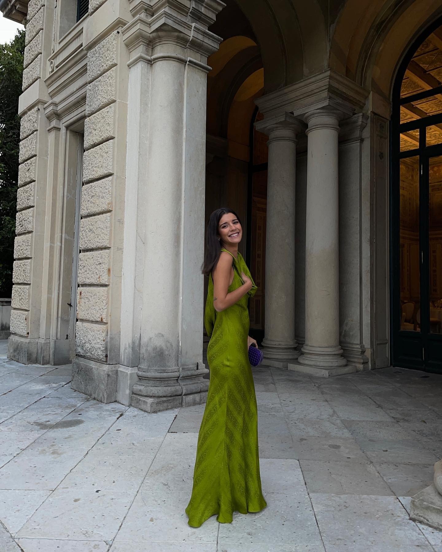 Look natal verde com vestido longo de tecido fluido e brilho sutil, modelo sorri confiante em cenário clássico e elegante.