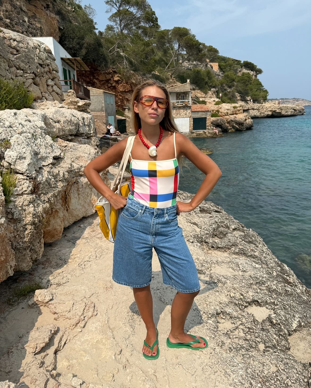 O look praia para o verão 2026 combina estilo e conforto, apresentando um top de biquíni listrado em cores vibrantes, acompanhado de shorts jeans. A modelo usa óculos de sol modernos e um colar com detalhes em concha, complementando a produção com uma