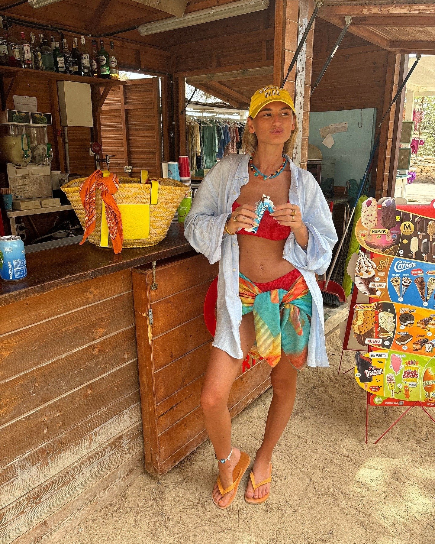 O look praia para o verão 2026 destaca um estilo descontraído e vibrante. A modelo usa um top vermelho, com um pareô colorido amarrado na cintura, que traz uma mistura de tons alegres.