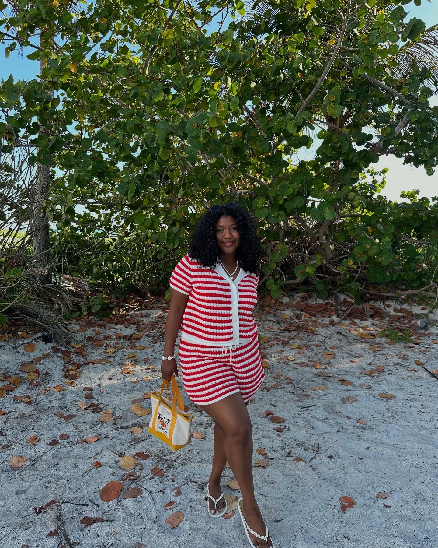 O look praia para o verão 2026 apresenta uma combinação vibrante de cores e conforto. A modelo usa um vestido curto listrado em vermelho e branco, ideal para dias ensolarados.