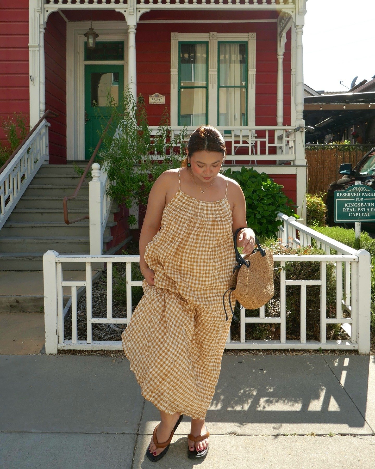 O look praia para o verão 2026 apresenta um vestido longo de gingham em tons de amarelo e branco, ideal para dias ensolarados. A modelagem leve e fluida proporciona conforto, enquanto a combinação com sandálias rasteiras e uma bolsa de palha complementa