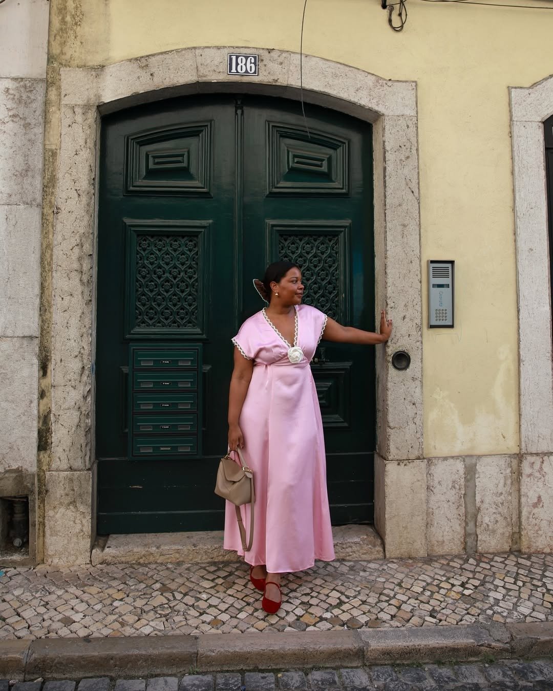 Mulher posa com look rosa em vestido longo de cetim com detalhes em renda e flor na cintura, combinando bolsa bege e sapato.