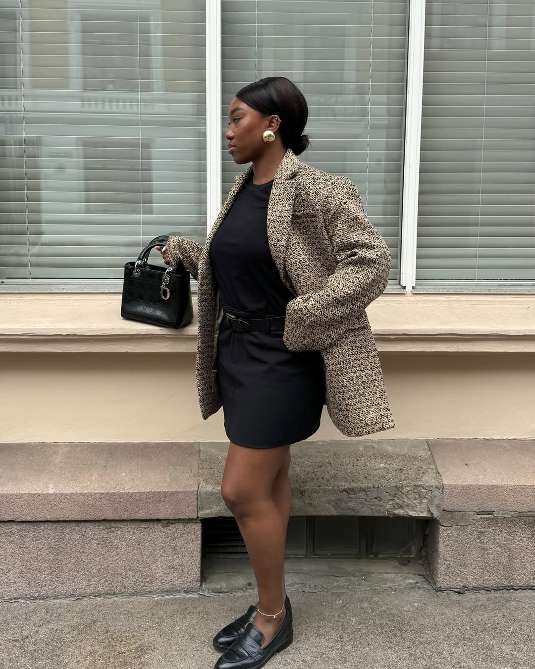 O look t-shirt é composto por uma camiseta preta básica, que combina perfeitamente com uma saia curta e um blazer texturizado em tons neutros. Os acessórios, como brincos grandes e uma bolsa preta, adicionam sofisticação.