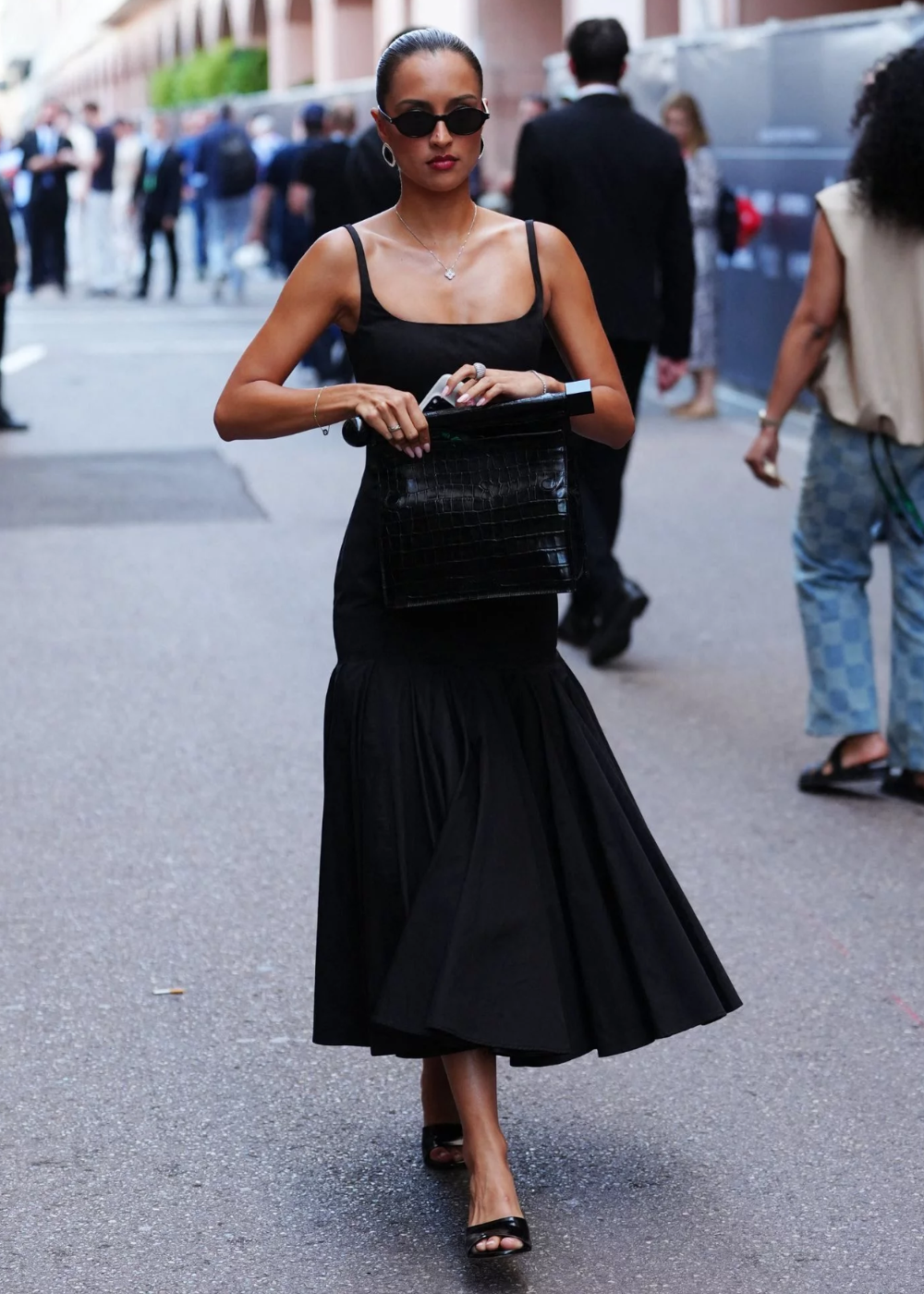 Alexandra Saint Mleux exibe um look sofisticado com um vestido preto de alças finas, que destaca sua silhueta elegante. Ela complementa a produção com acessórios minimalistas, como óculos escuros e brincos discretos, além de uma bolsa de couro croco.