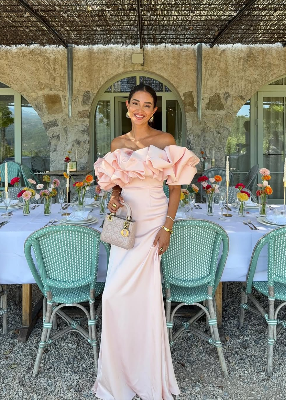 Modelo sorri confiante posando com vestido longo rosa claro de tecido fluido e babados volumosos ombro a ombro, look black