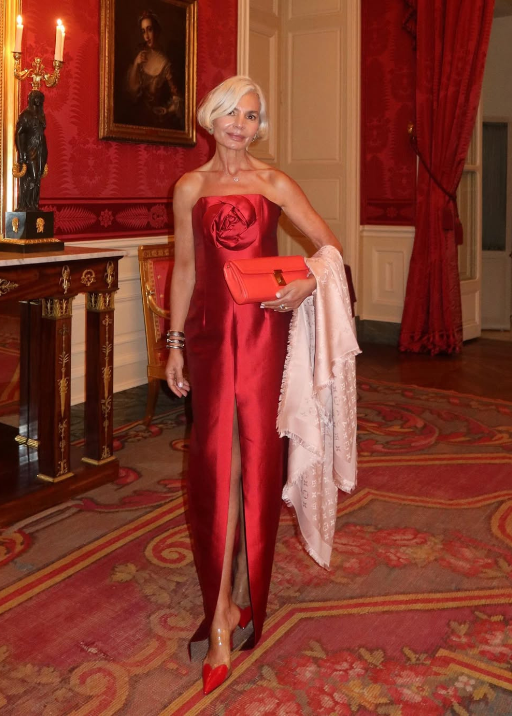 Mulher posa com look black tie em vestido vermelho acetinado tomara que caia com fenda frontal, clutch coral e lenço rosa