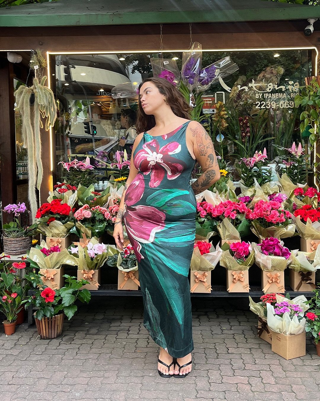Modelo posa em vestido tendência longo justo de tecido leve com estampa floral grande em tons de verde, rosa e vermelho, em