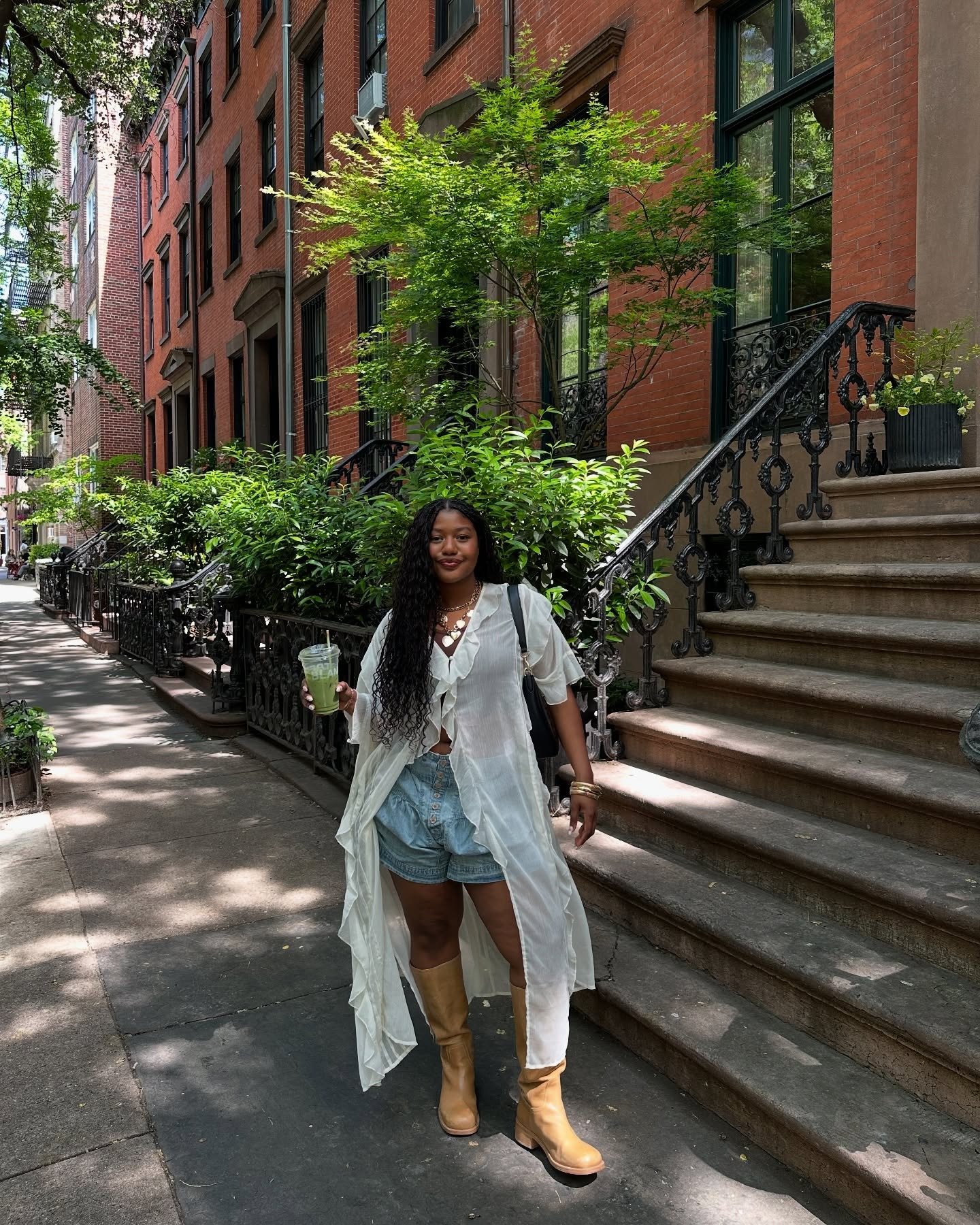 Modelo posa na rua com look casual de shorts jeans, camisa branca longa e fluida e bota cano alto caramelo em couro, clima