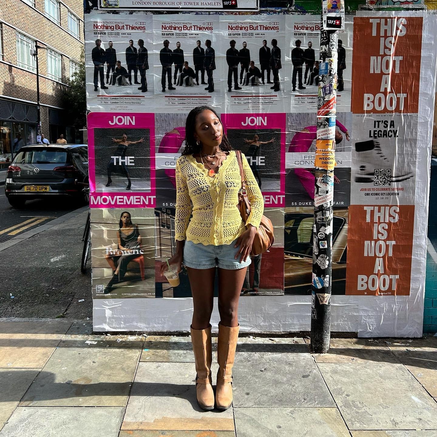 Modelo posa com looks com bota cano alto bege em couro, short jeans claro e blusa amarela de crochê, em clima urbano e