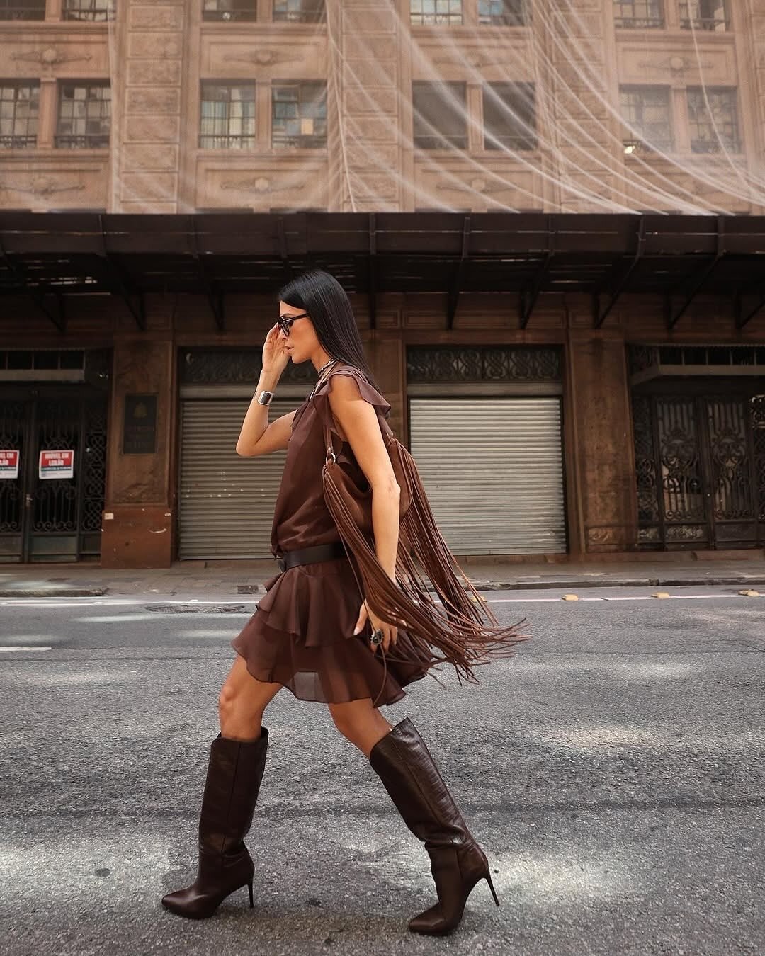 Mulher usa veste marrom com bolsas de franjas e bota de cano alto com salto fino.