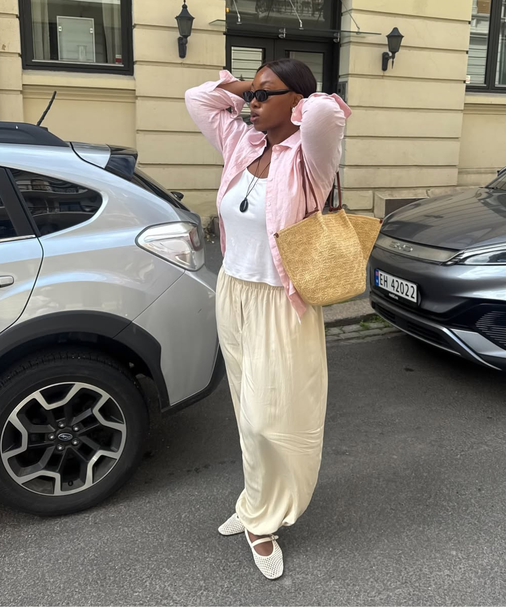 Modelo posa com looks com calça balonê em tecido fluido creme, combinada a camiseta branca, camisa rosa e bolsa de palha, em