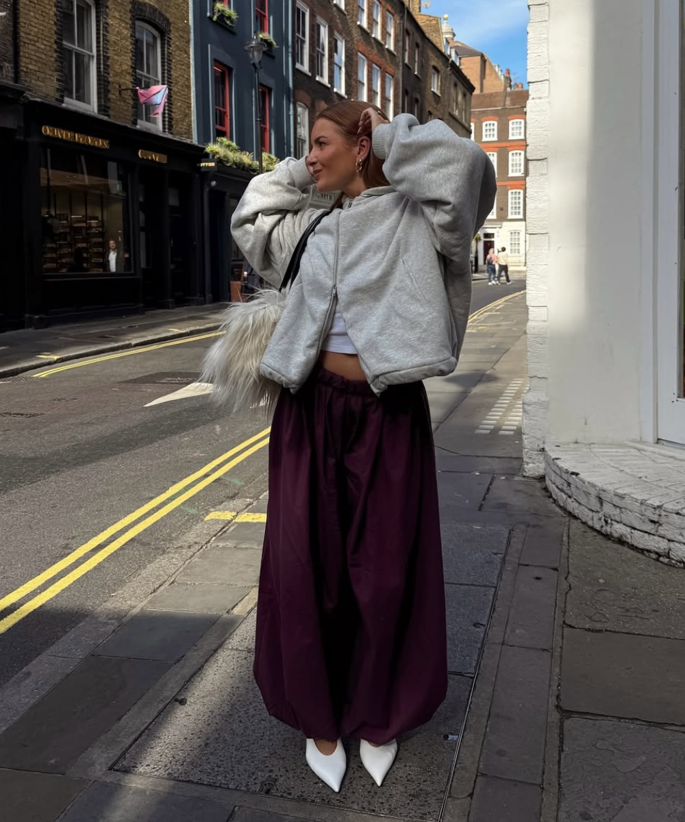 Modelo posa com look urbano que destaca calça balonê vinho de tecido leve, moletom cinza oversized e scarpin branco,