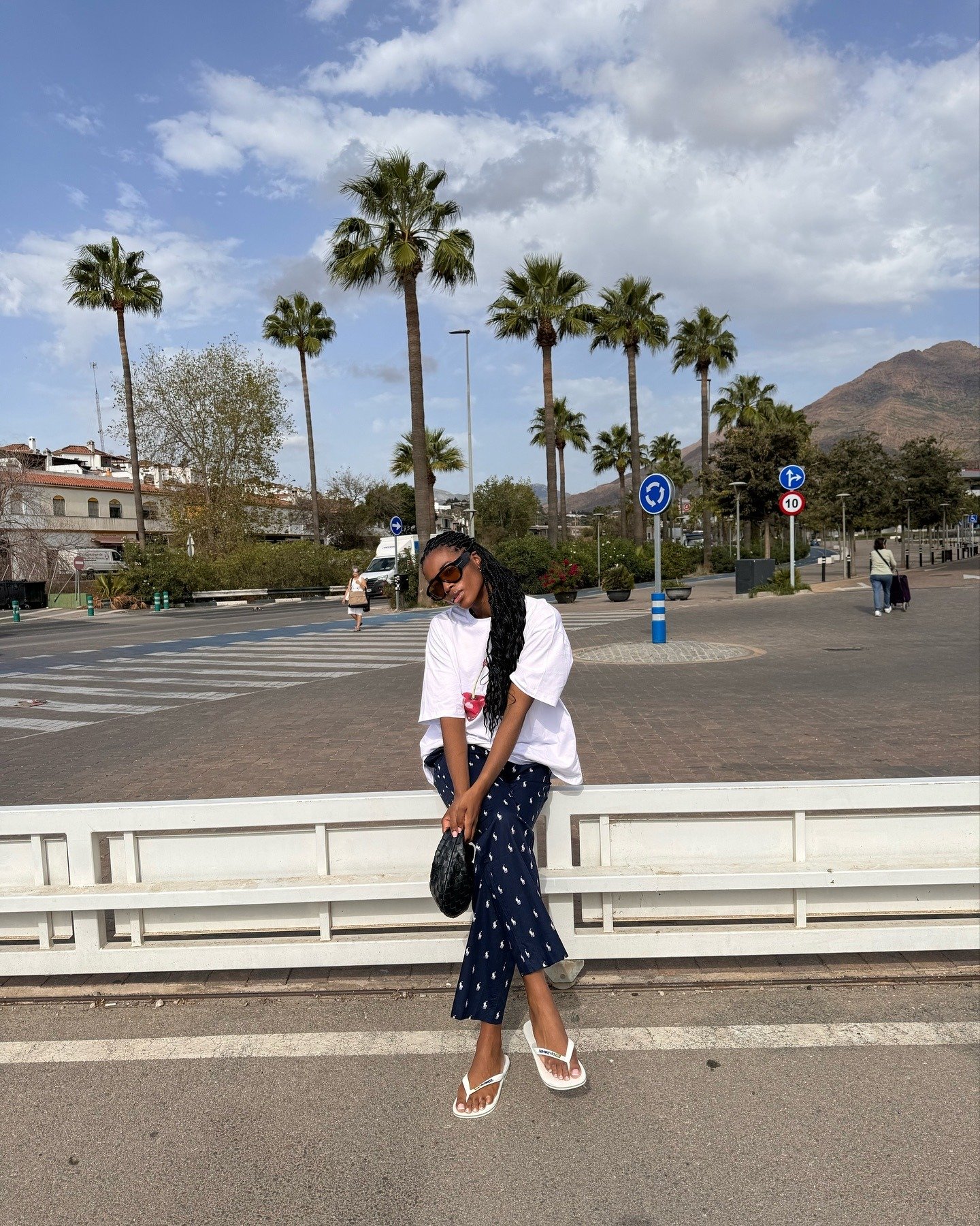 Modelo senta casualmente em banco urbano usando chinelo branco de tiras finas, camiseta branca ampla e calça azul estampada,