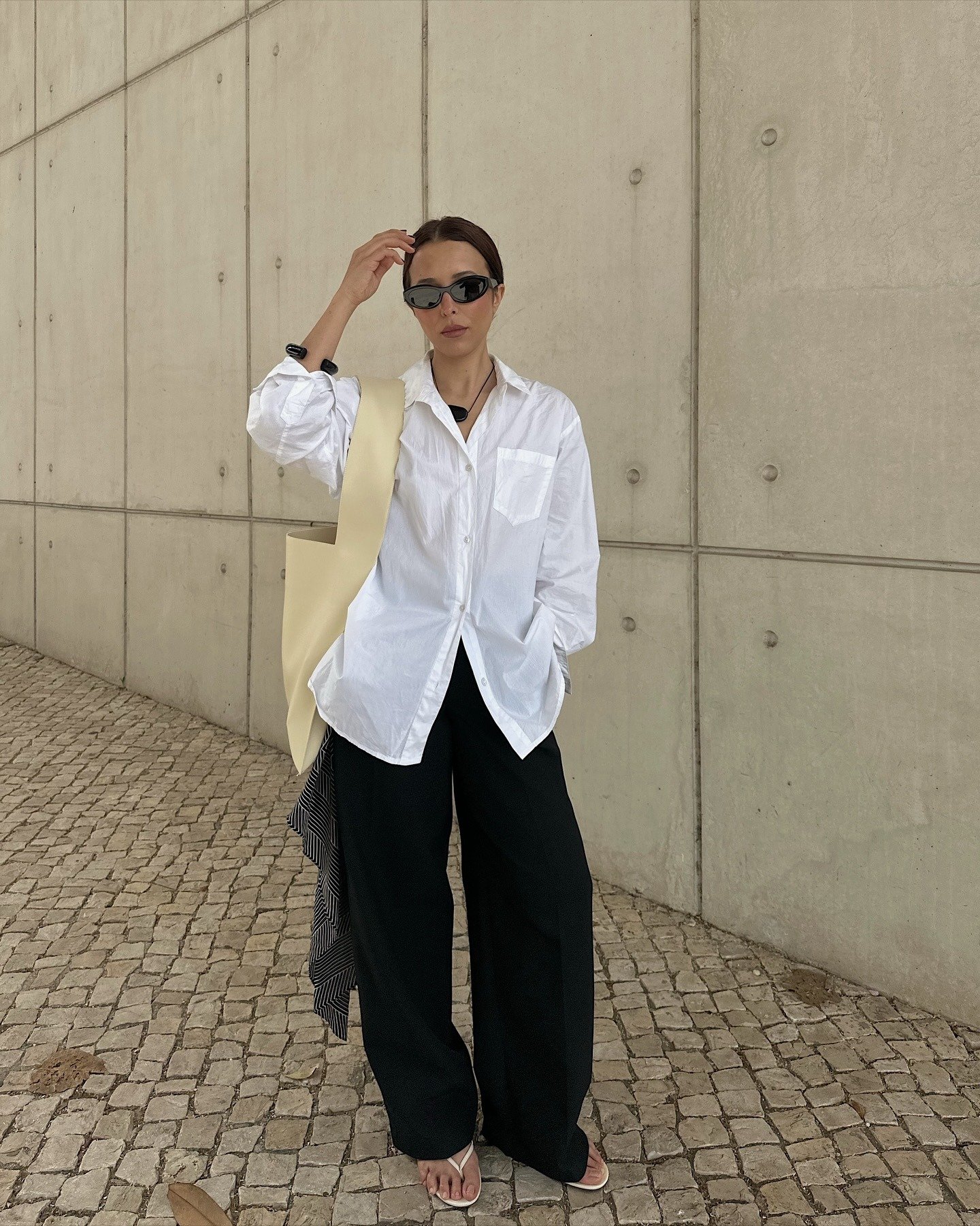 Modelo posa com camisa branca oversized, calça preta ampla e chinelo branco minimalista, em clima urbano e sofisticado.