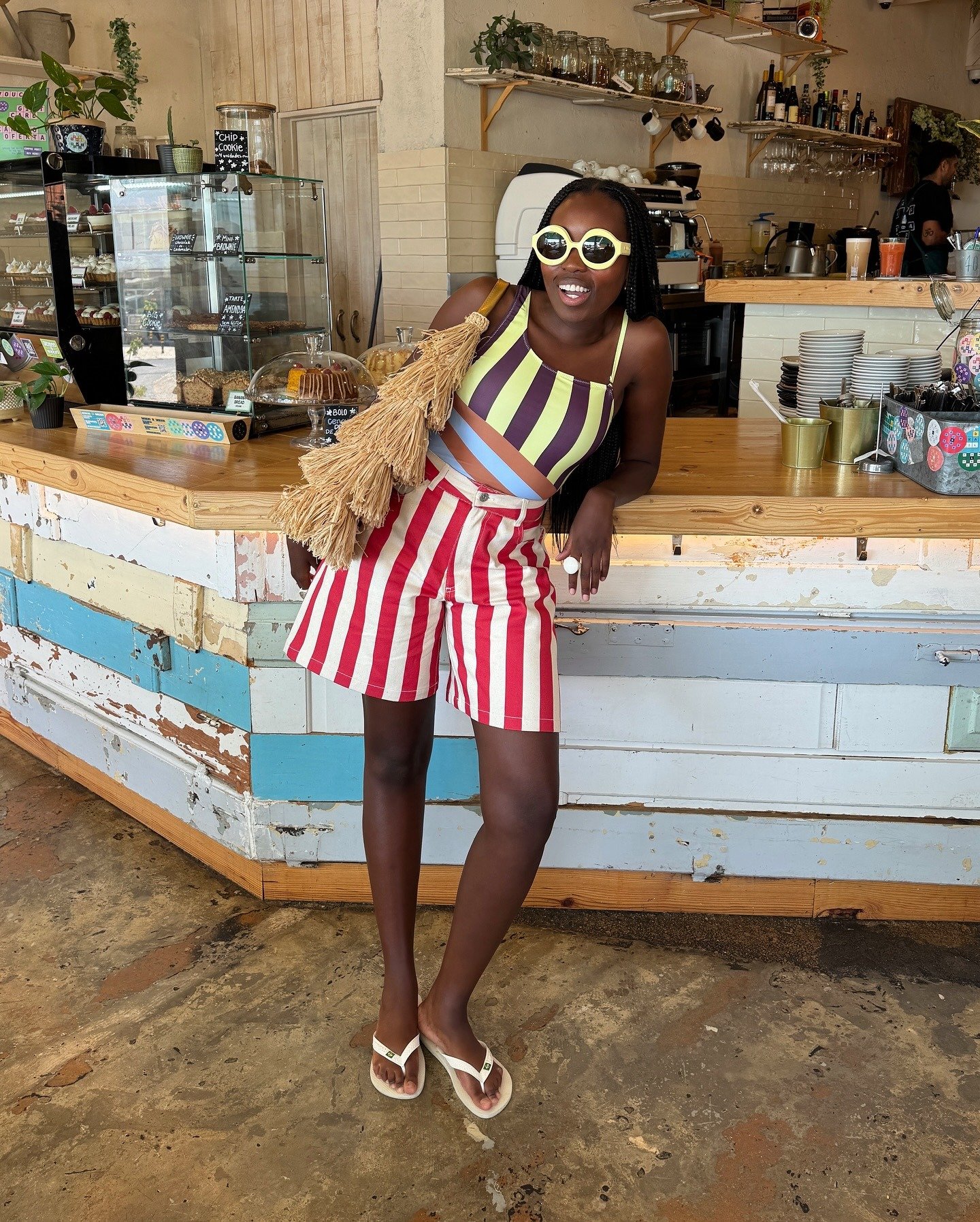 Modelo posa sorridente apoiada no balcão rústico usando chinelo branco, short listrado vermelho e branco, top colorido e
