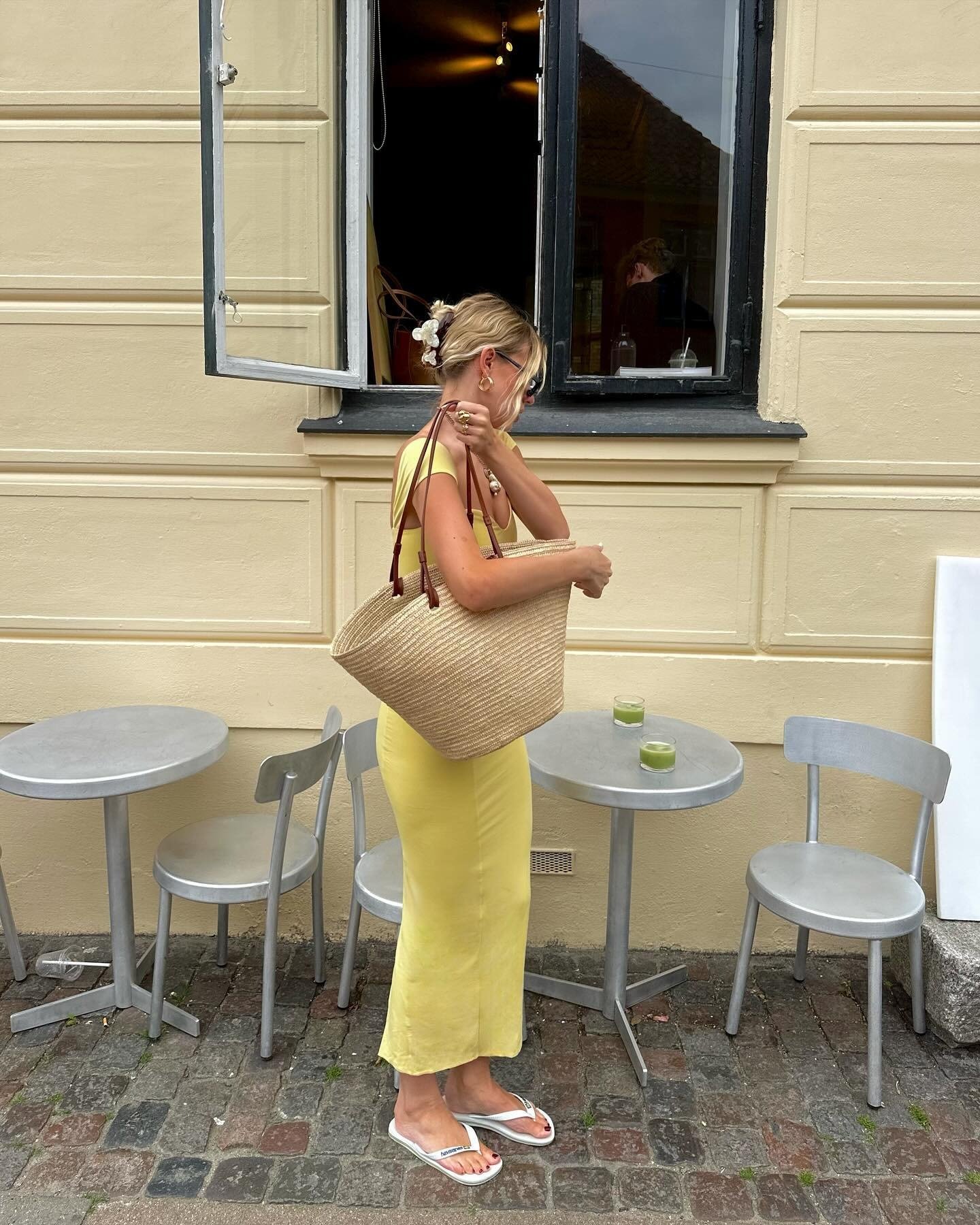 Mulher em vestido amarelo justo de tecido leve usa chinelo branco simples, bolsa de palha e acessórios dourados, em clima