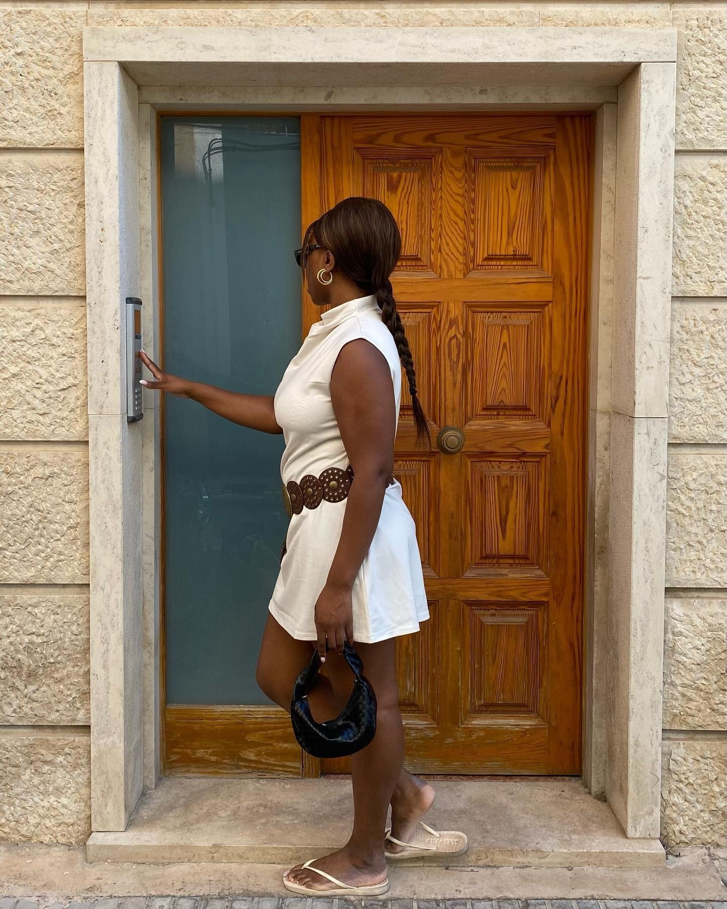 Mulher em vestido branco curto com cinto marrom larga, chinelo branco minimalista e bolsa preta pequena, em postura casual