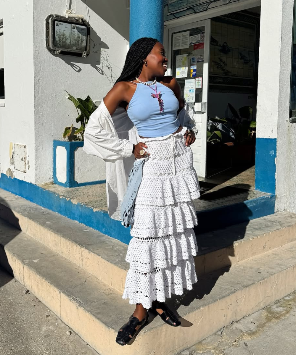 Modelo sorri e posa com look com crochê composto por saia branca de babados, regata azul e camisa branca leve, transmitindo
