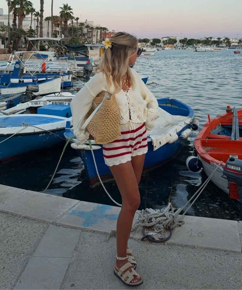 Modelo posa de perfil com look com crochê composto por cardigan branco de manga longa, shorts listrados vermelhos e brancos,