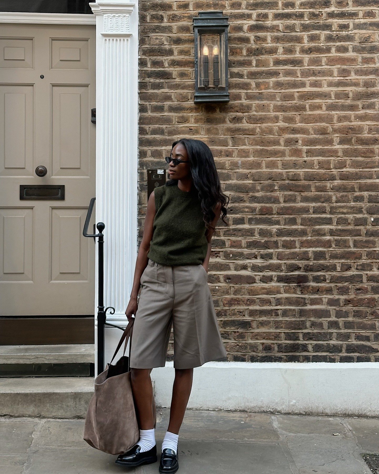 Este looks com loafer de couro apresenta uma combinação sofisticada e casual. A blusa verde-oliva é complementada por bermudas bege de corte reto, criando um visual equilibrado. O uso de meias brancas e a bolsa de camurça adicionam um toque moderno.