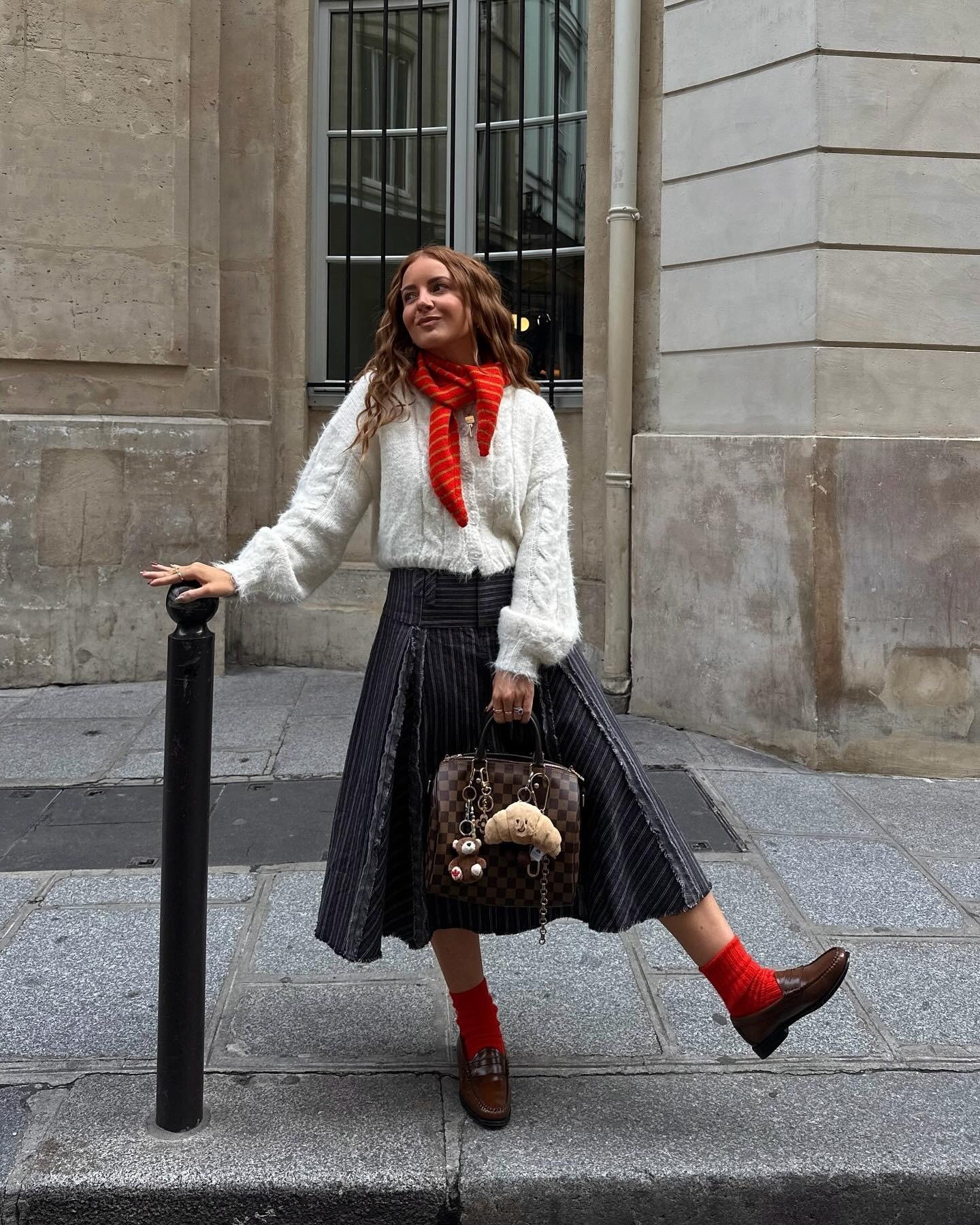 O look com loafer de couro destaca-se pela combinação de um suéter felpudo branco e uma saia midi listrada, criando um contraste elegante. O toque vibrante do lenço vermelho e das meias combinando adiciona personalidade.