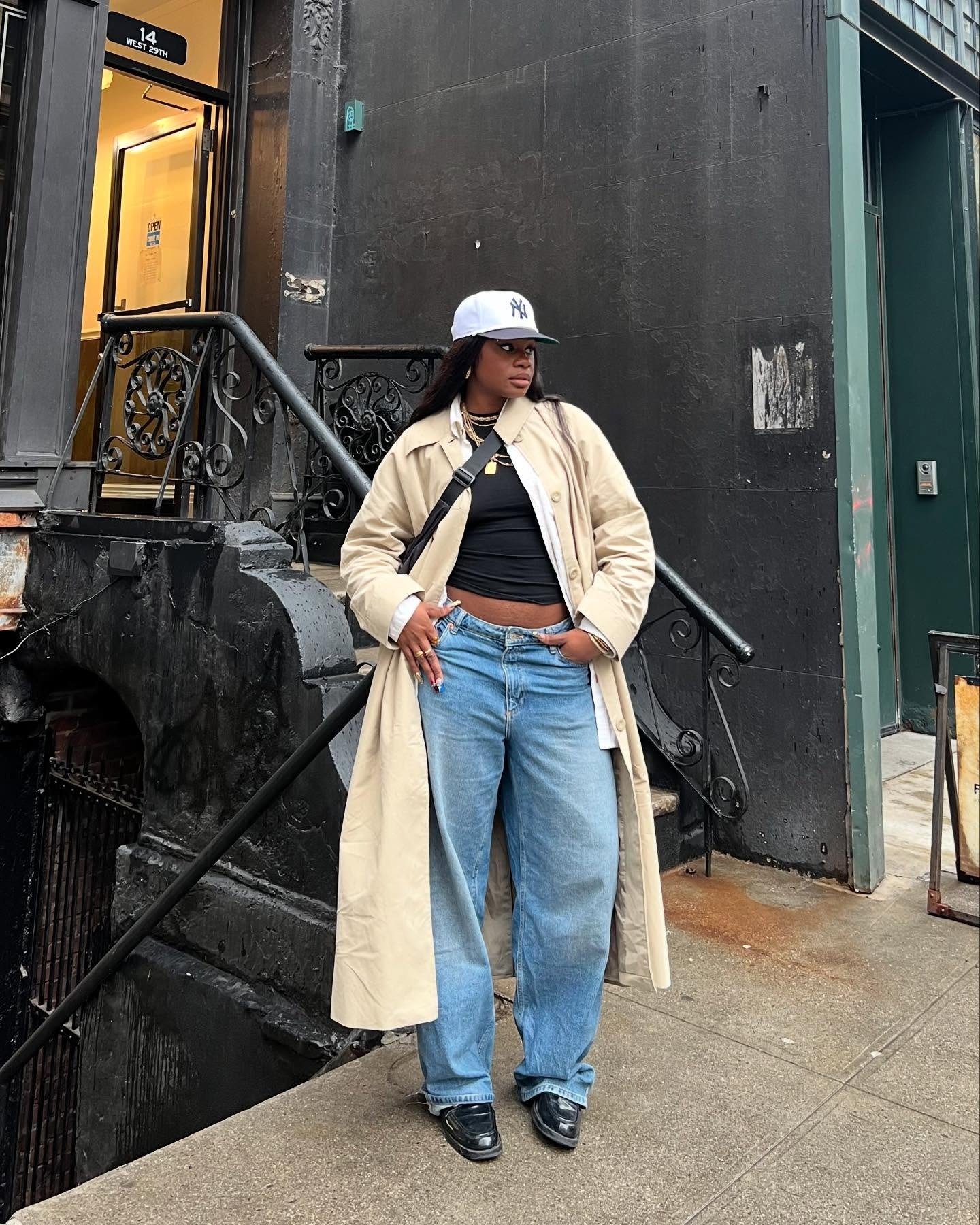 O look apresenta uma combinação estilosa com loafer de couro preto, que confere elegância ao visual. A modelo usa um trench coat bege sobre uma blusa preta, combinando com jeans de corte amplo. O boné branco adiciona um toque casual.