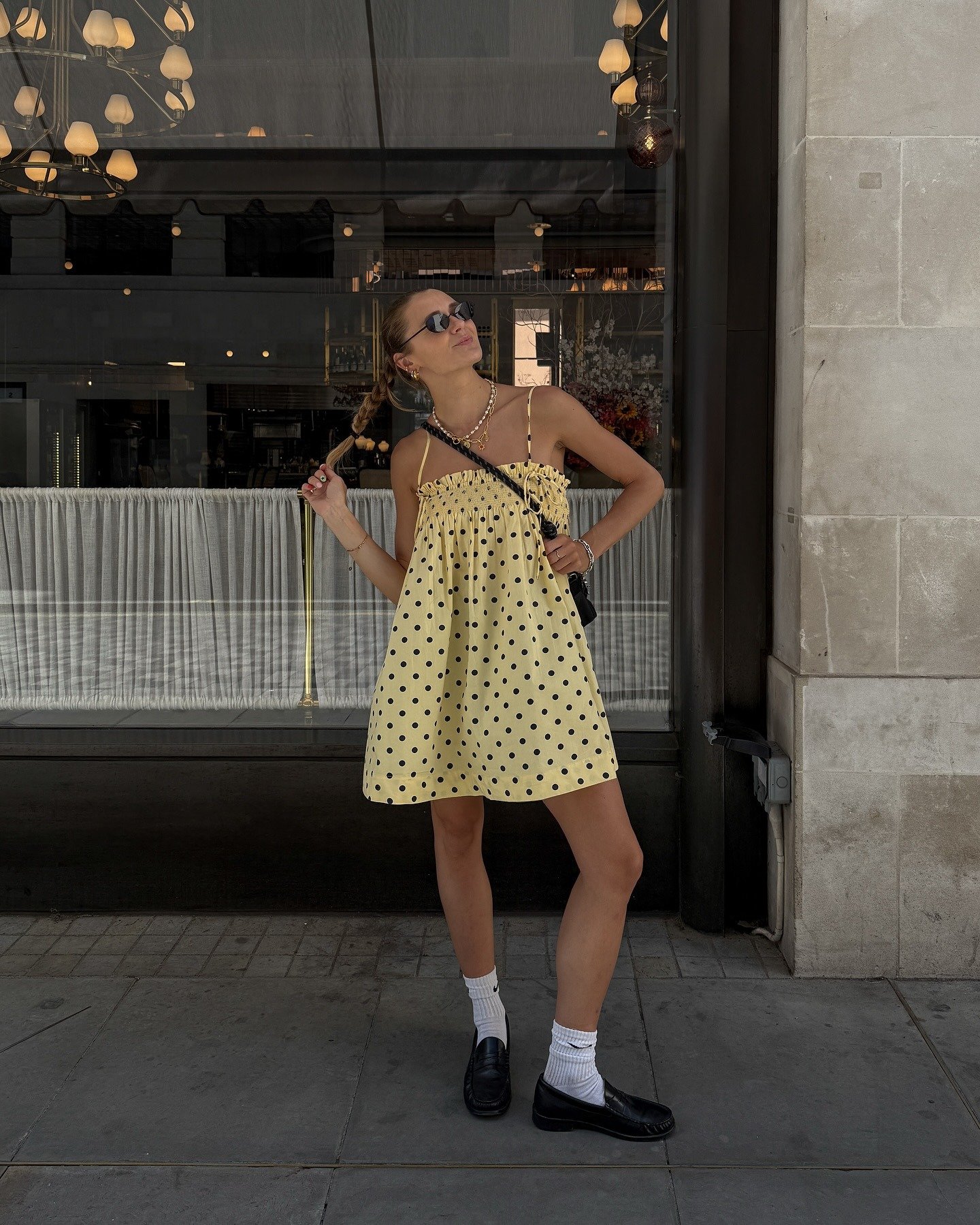 O look destaca um vestido amarelo com poás pretos, ideal para dias ensolarados. A combinação com loafers de couro pretos traz um toque de sofisticação e conforto. Meias brancas adicionam um contraste interessante, equilibrando a leveza do vestido.