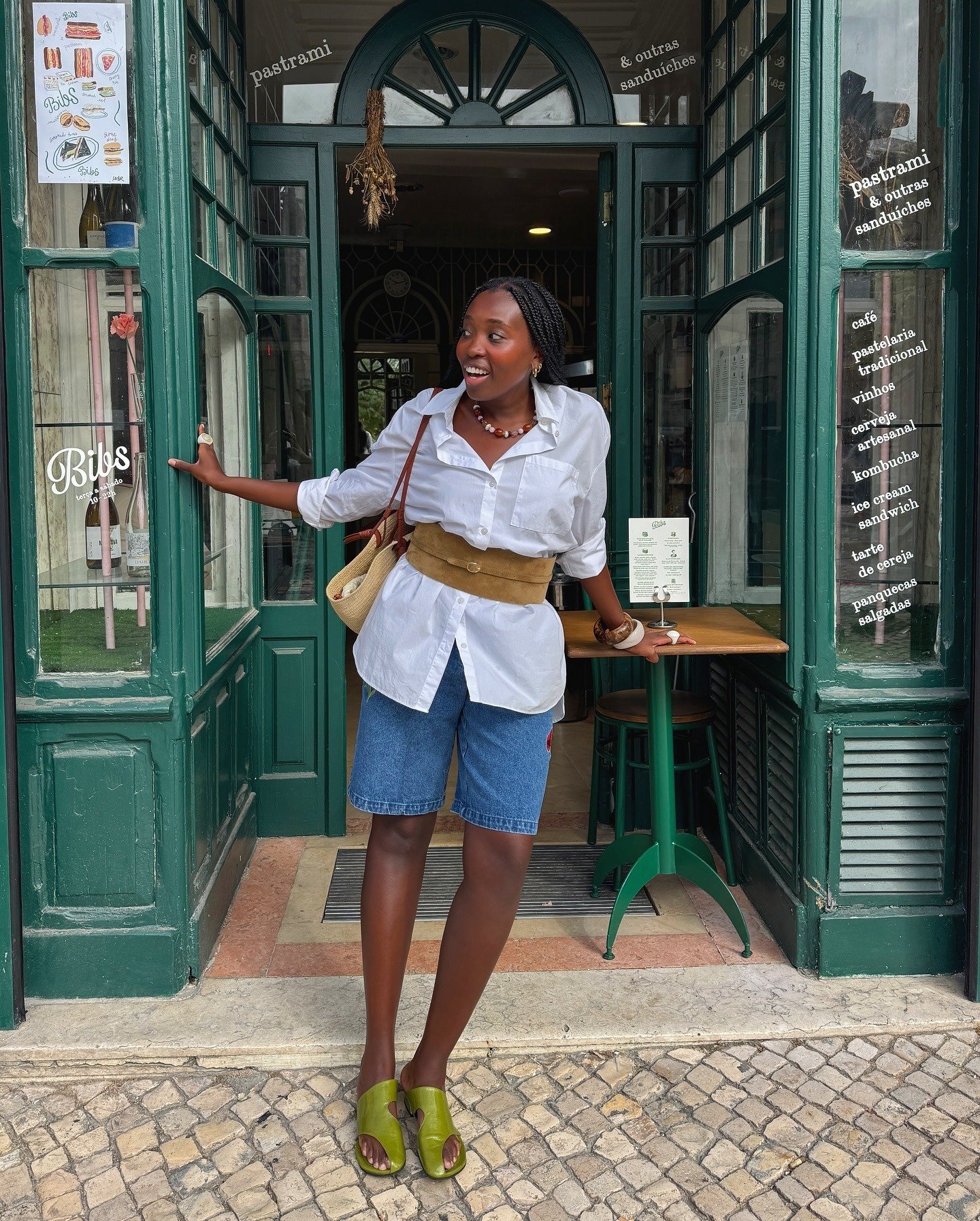 Modelo posa sorrindo na entrada de café com camisa branca oversized, bermuda jeans, cinto largo caramelo e rasteirinha