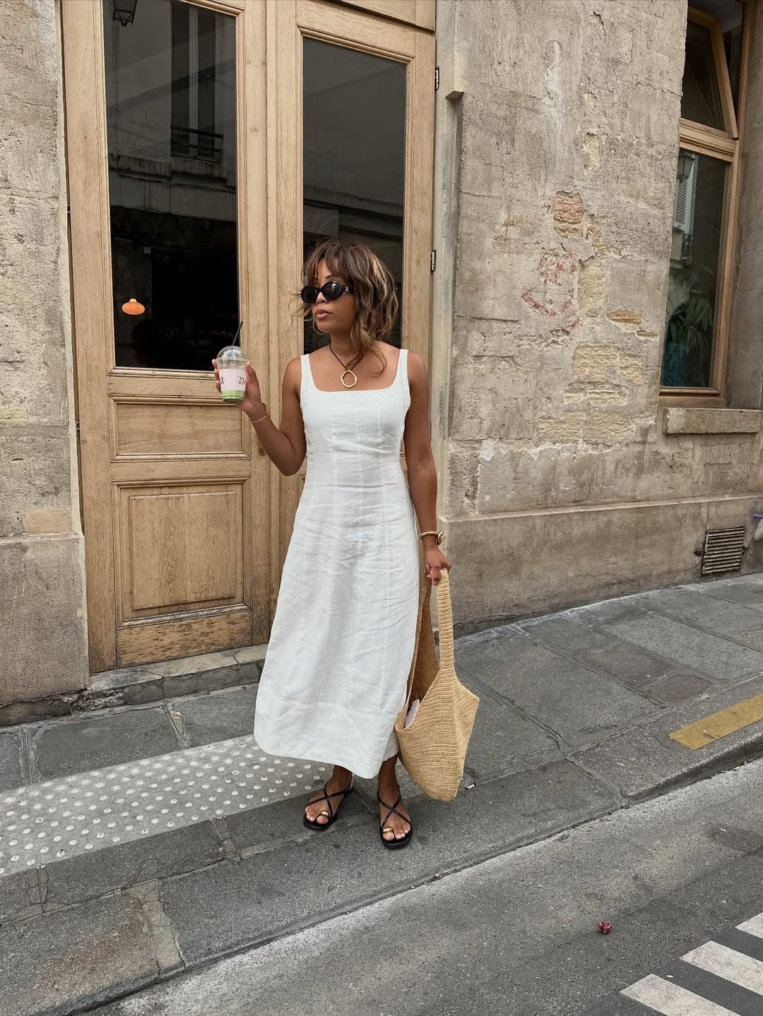 Mulher posa com vestido midi branco de linho, rasteirinha preta de tiras finas e bolsa de palha, em clima casual e elegante.