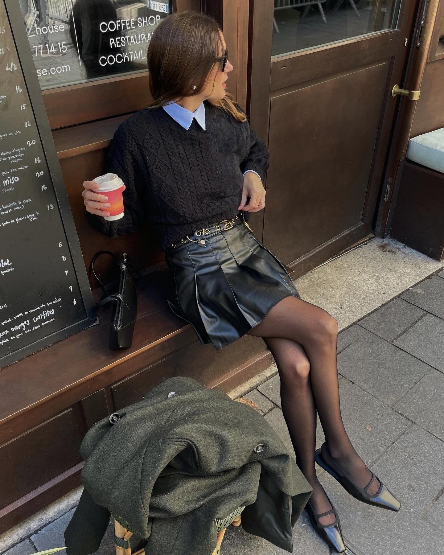 O visual apresenta uma saia preta curta de couro, combinada com um suéter preto de tricô e uma camisa com colarinho branco. Acessórios como um cinto dourado e óculos escuros complementam o look.