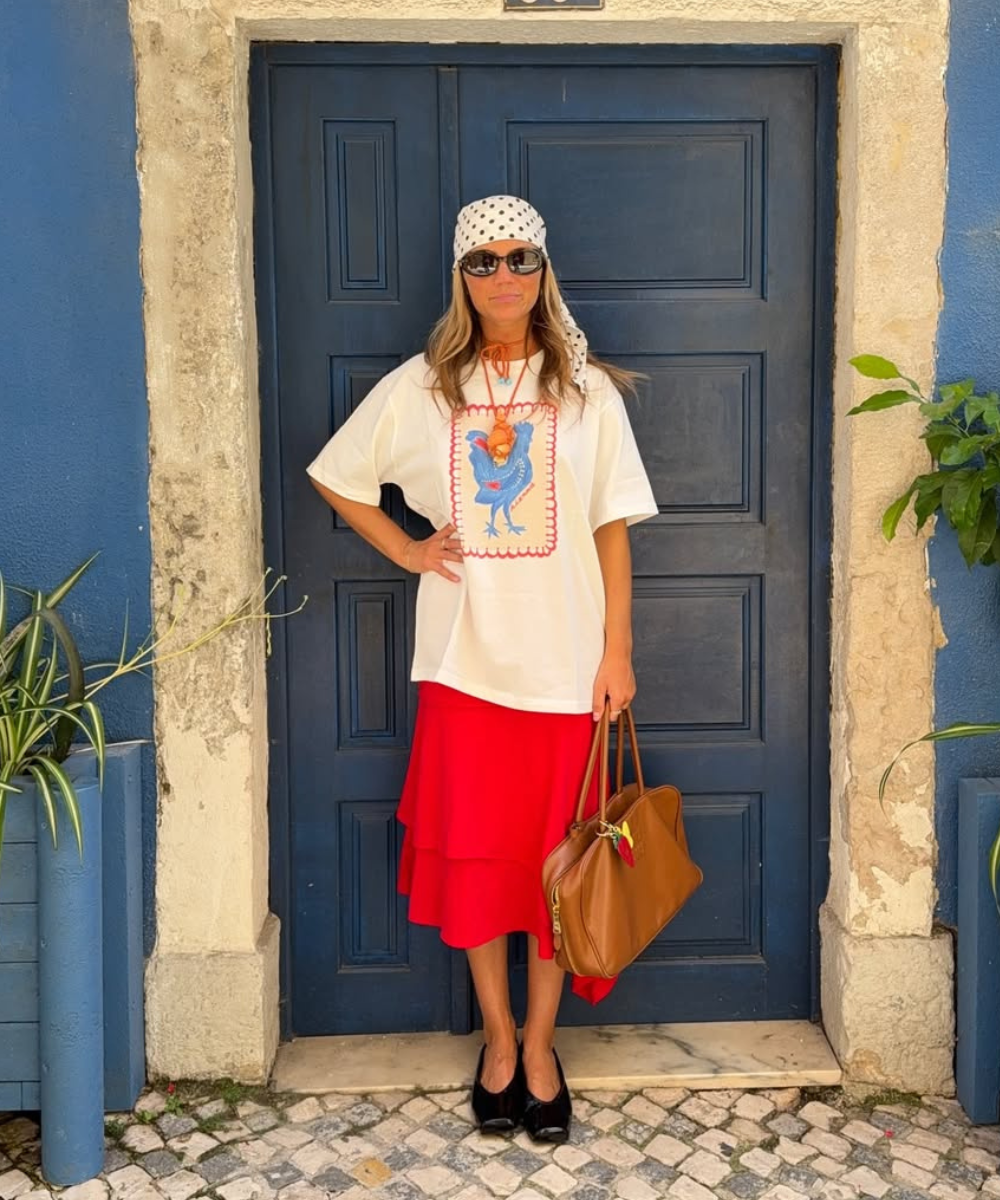 Modelo posa com look casual de saia vermelha em camadas, camiseta branca estampada, lenço de poás e bolsa caramelo, estilo.