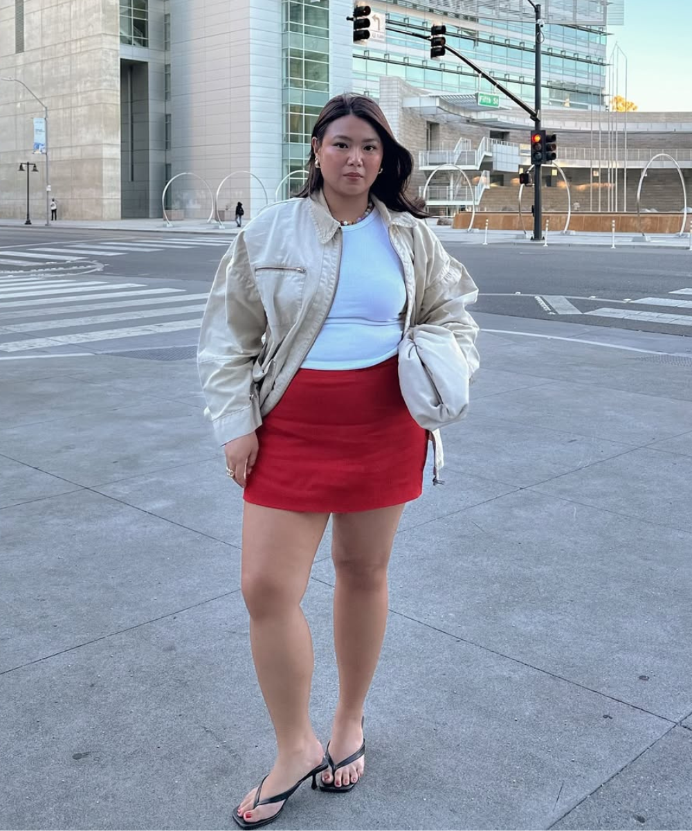 Look com saia vermelha curta em tecido liso, combinada a regata branca e jaqueta bege oversized, estilo casual urbano.