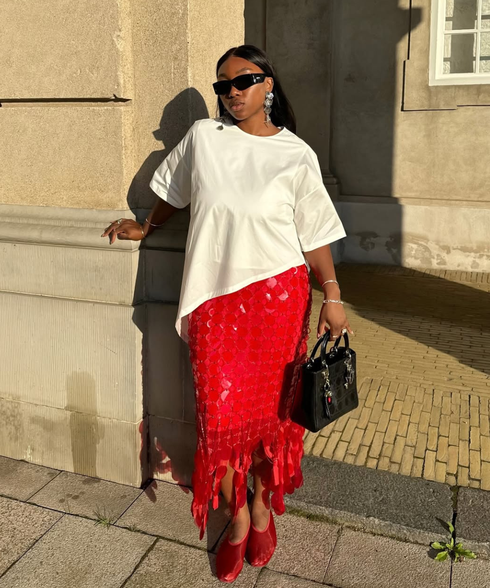 Modelo posa com look moderno em saia vermelha midi de textura vazada e babados, combinada a blusa branca assimétrica e