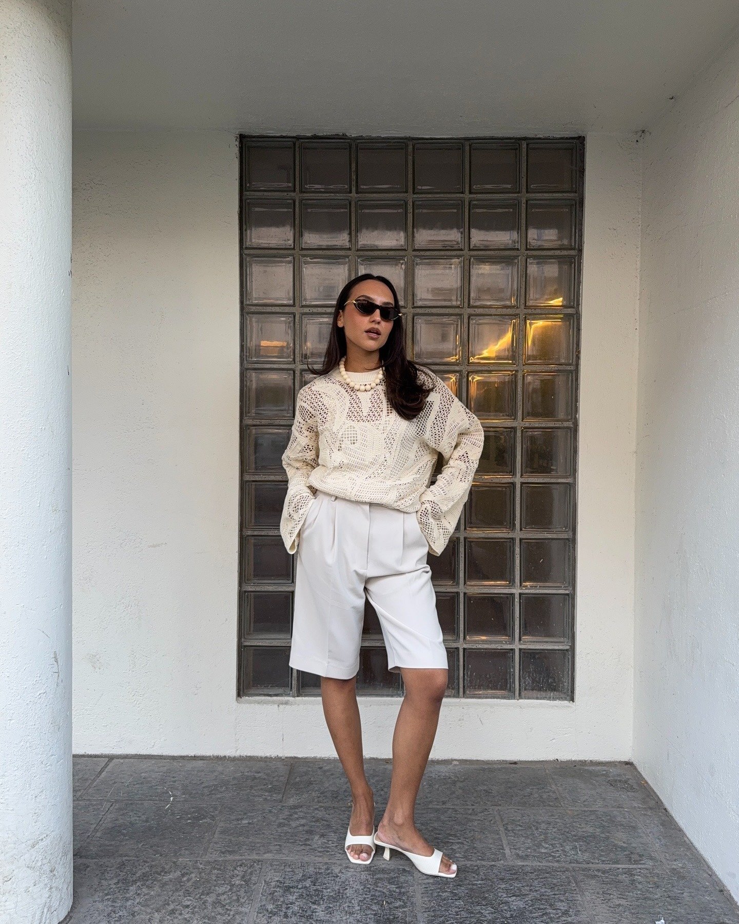 Look com sandália branca de salto baixo, bermuda alfaiataria e blusa de tricô vazada, estilo casual chic com postura