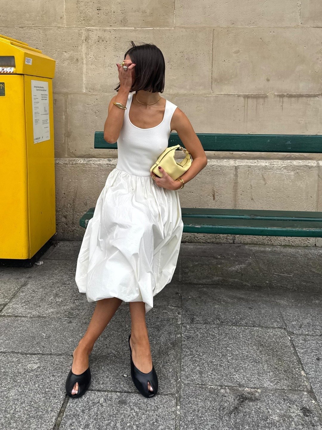 A imagem apresenta uma mulher sentada em um banco, vestindo um vestido branco com saia volumosa e uma blusa de alças. Ela segura uma bolsa amarela e usa sapatos peep-toe pretos, que destacam seus pés e adicionam um toque elegante ao visual.