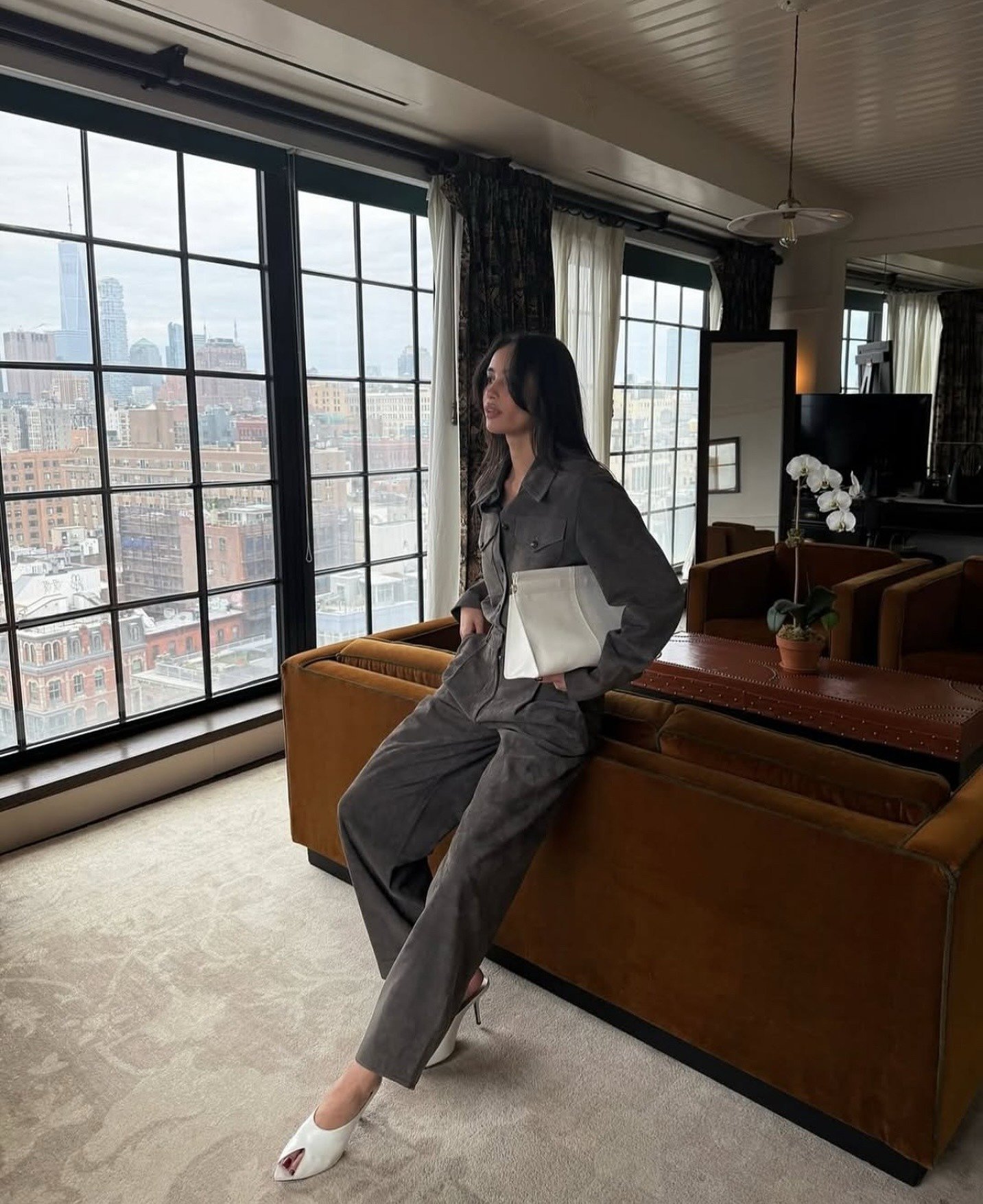 A imagem apresenta uma mulher em um ambiente sofisticado, usando um conjunto de alfaiataria em tom cinza. Nos pés, destaca-se um elegante sapato peep-toe branco, que revela os dedos e adiciona um toque de modernidade ao look.