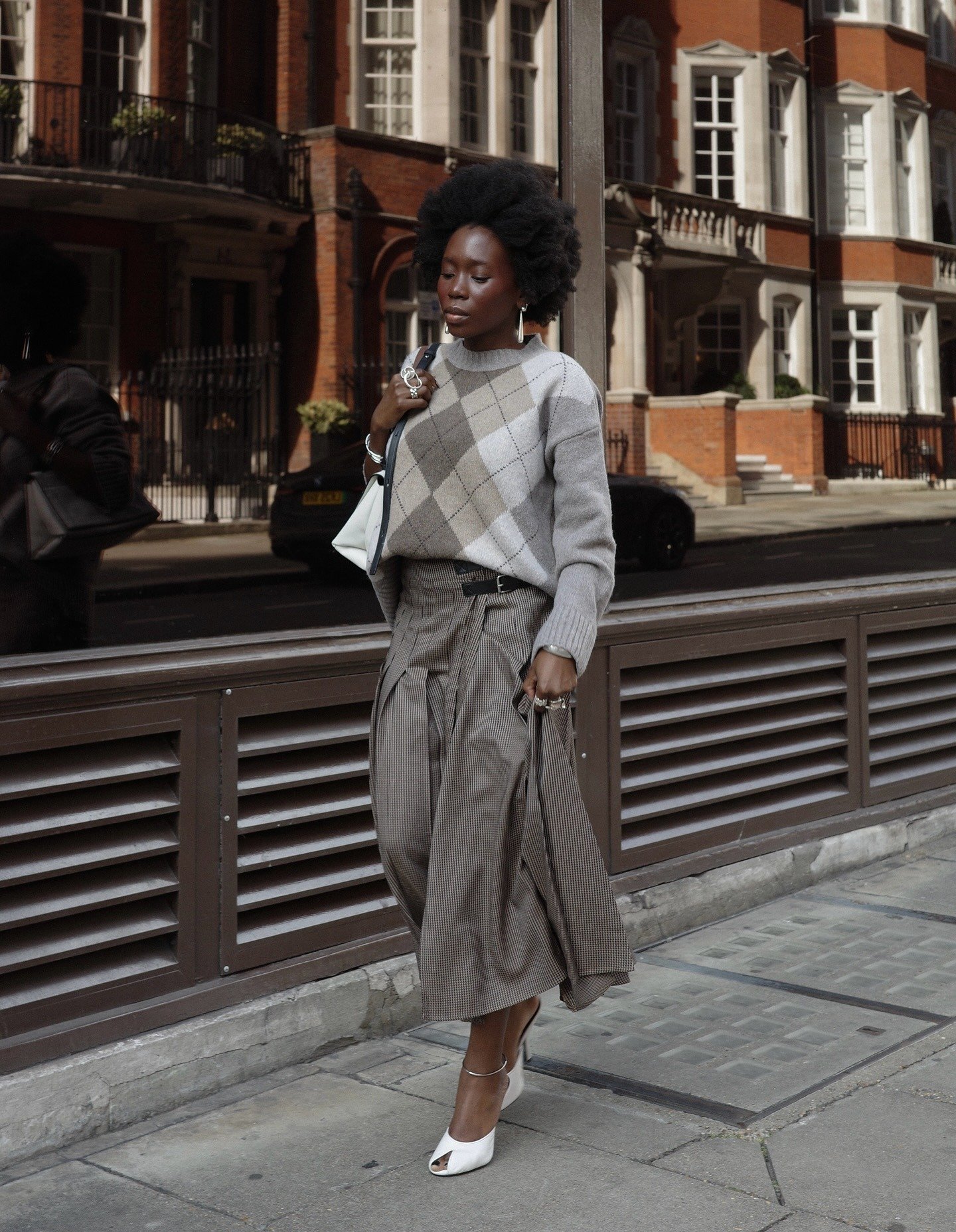 O sapato peep-toe em destaque combina elegância e modernidade, perfeito para compor looks sofisticados. A modelo, vestindo um suéter de padrão argyle e uma saia midi, exibe o calçado branco, que revela os dedos dos pés e adiciona um toque contemporâneo