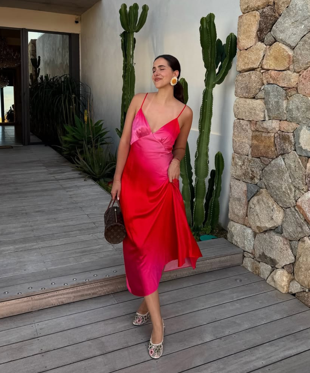 O look apresenta um vestido vermelho em gradiente, com alças finas e um caimento fluido, ideal para festas de Natal. A modelo complementa a produção com brincos grandes e uma bolsa elegante.