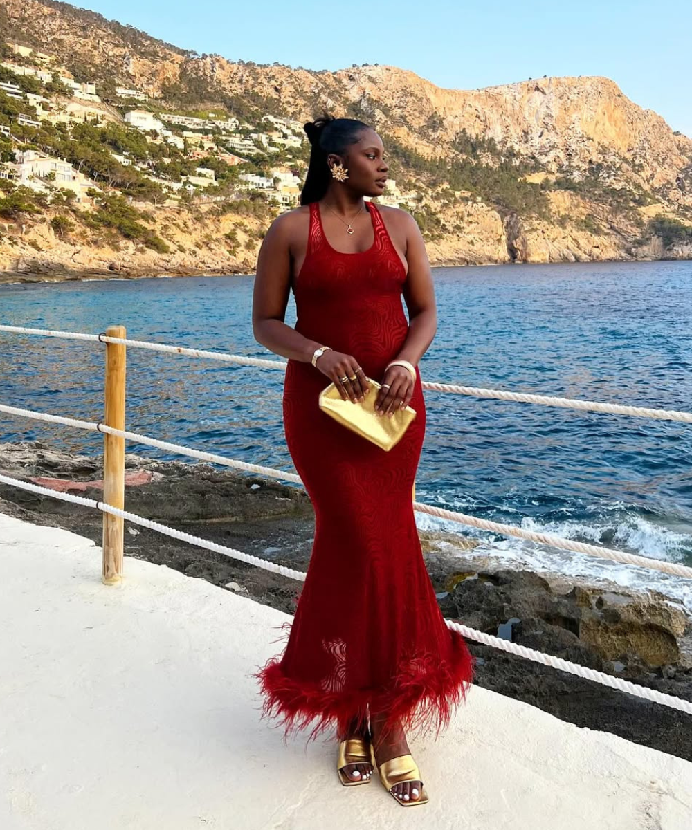 O look apresenta um vestido vermelho justo, com detalhes em renda e um acabamento com plumas na barra, ideal para festas de Natal. A combinação com acessórios dourados, como uma bolsa e brincos, traz sofisticação.