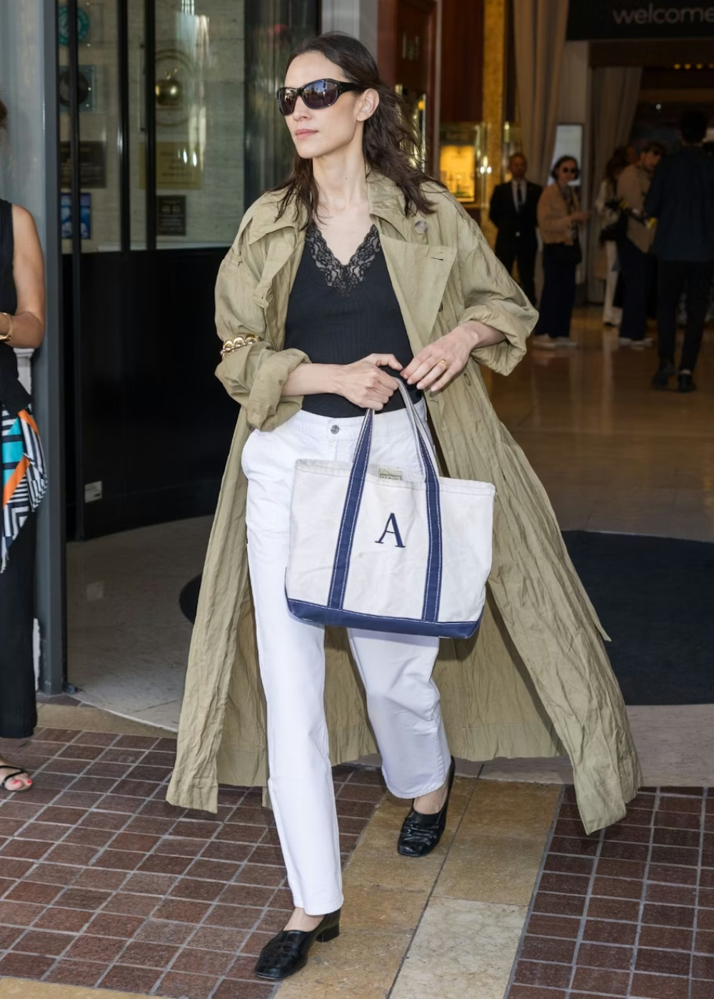 Alexa Chung caminha confiante com trench coat bege oversized, regata preta com renda, calça branca reta e bolsa branca com