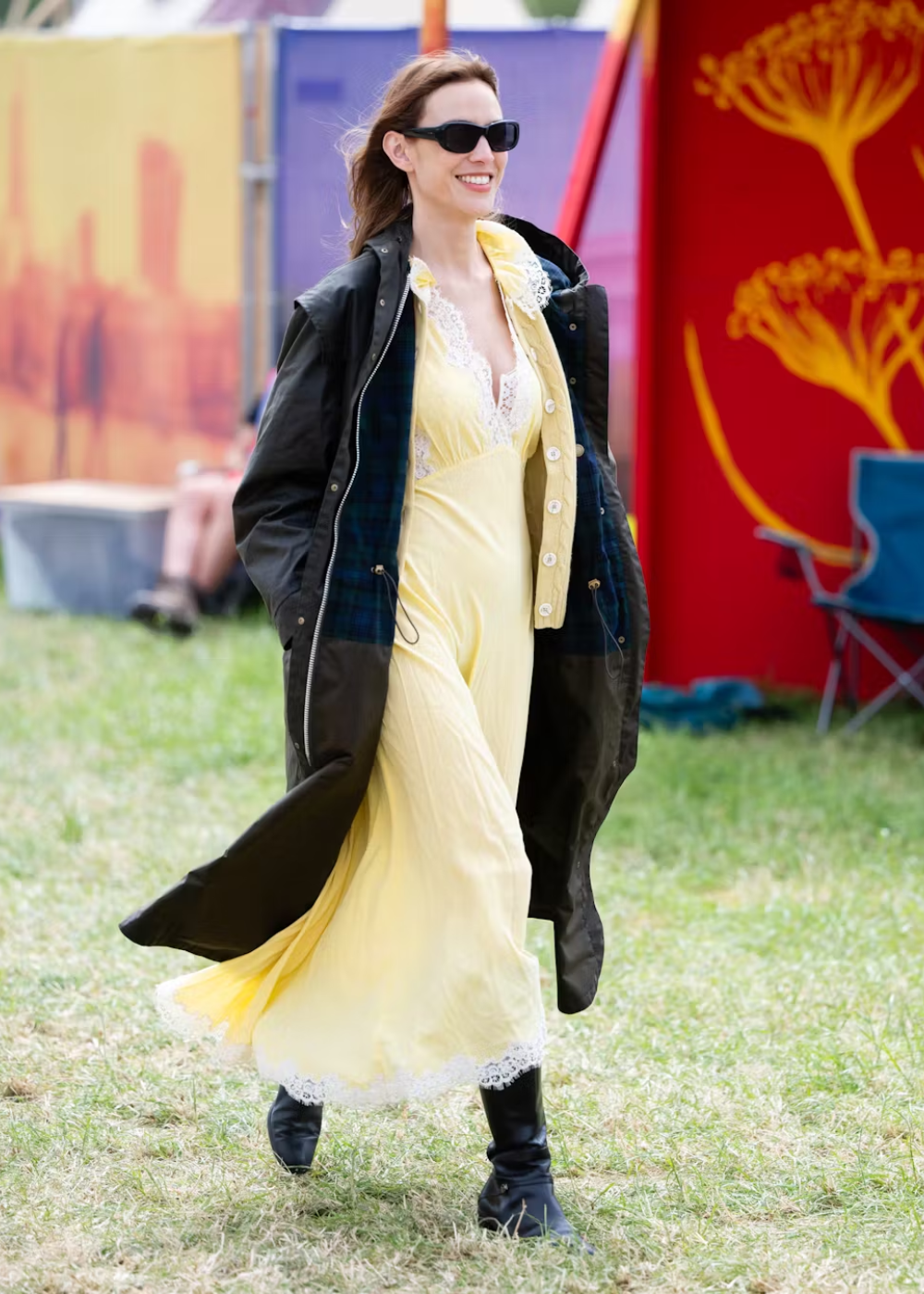 Alexa Chung caminha sorridente com vestido amarelo fluido de renda branca e maxi casaco preto, combinando botas pretas e